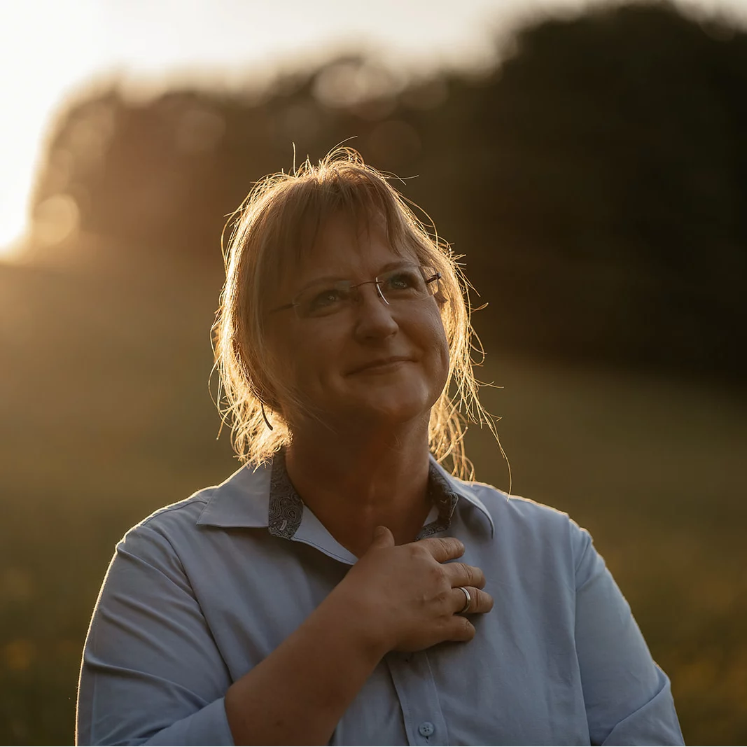 Person mit schulterlangem, hellbraunem Haar trägt hellblaues Hemd und Ring, Hand auf Brust, im Gegenlicht im Freien.