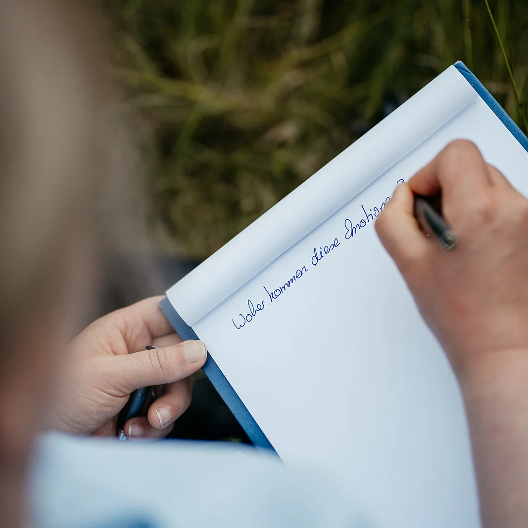 Person schreibt mit schwarzem Stift auf einem weißen Notizblock, Text beginnt mit 'Woher kommen diese Emoitionen'.