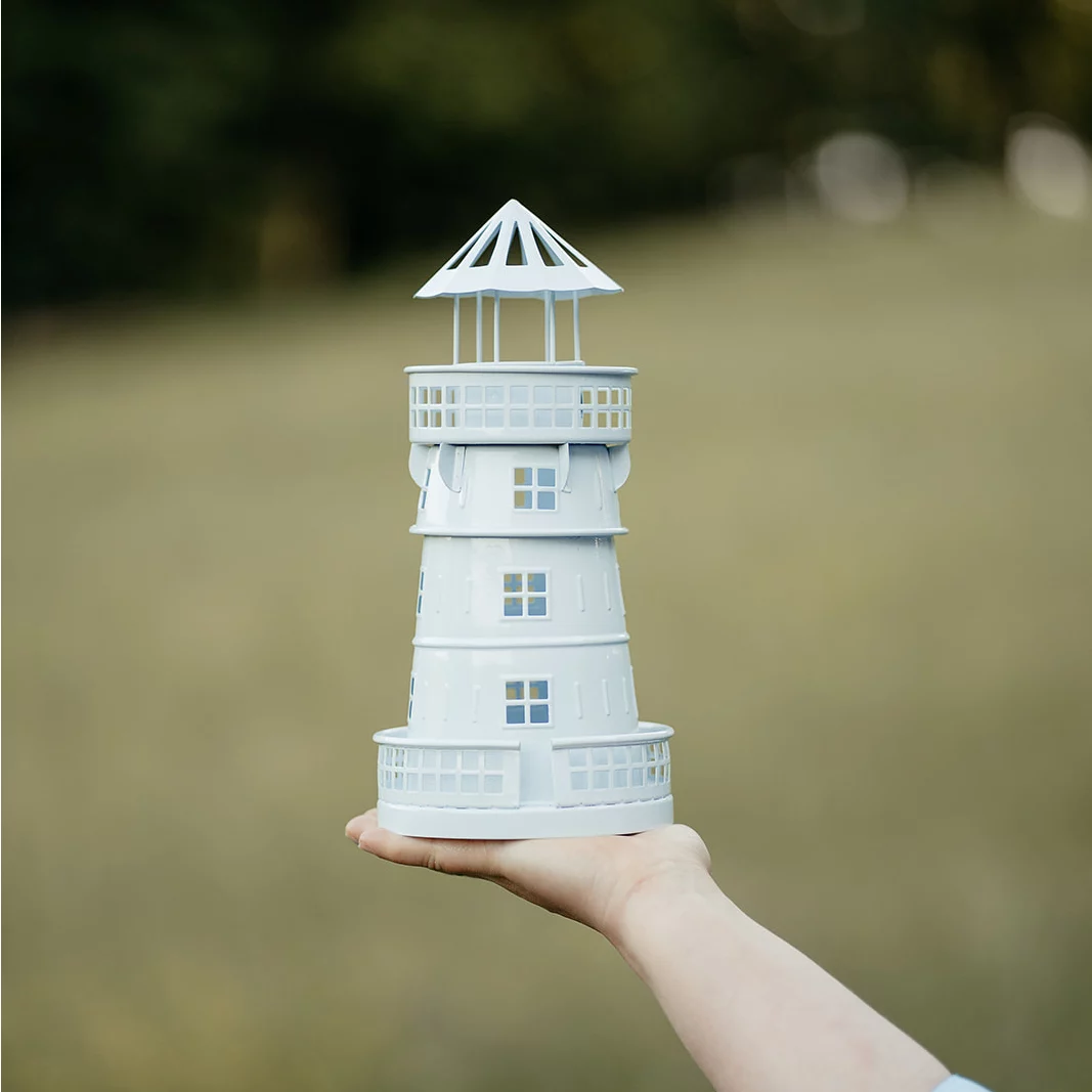 Hand hält ein weißes Modell eines Leuchtturms mit mehreren Fenstern und einer offenen Spitze vor unscharfem Hintergrund.
