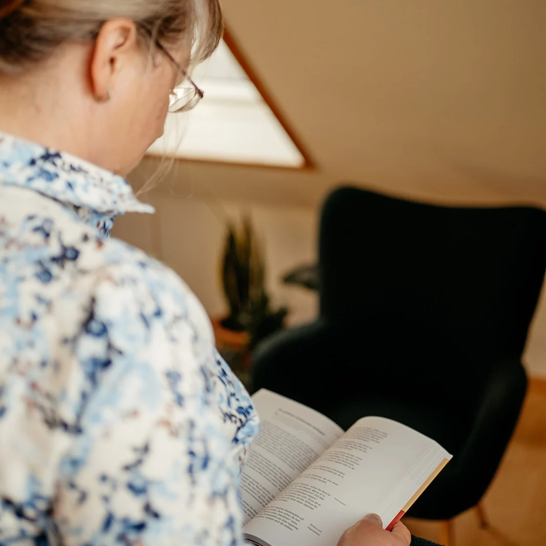 Frau mit Brille und gemustertem Hemd liest ein Buch vor einem schwarzen Sessel in einem Raum mit Dachschräge