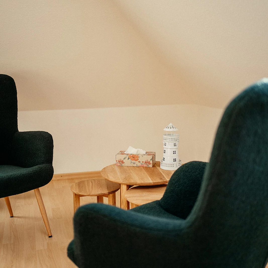 Zwei dunkelgrüne Sessel mit Holzbeinen vor einer beige gestrichenen Wand, dazwischen runde Holztische mit einer Papiertaschentuchbox und einem weißen Leuchtturm-Modell.